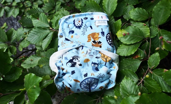 A cloth diaper with a blue background featuring illustrations of woodland animals such as a raccoon and an owl. The diaper is laying on a bed of lush green leaves.