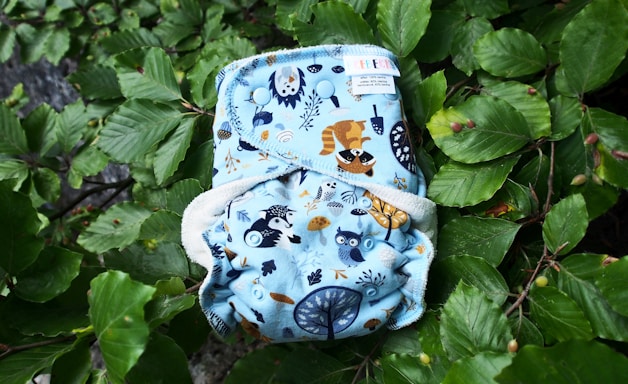 A cloth diaper with a blue background featuring illustrations of woodland animals such as a raccoon and an owl. The diaper is laying on a bed of lush green leaves.