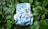 A cloth diaper with a blue background featuring illustrations of woodland animals such as a raccoon and an owl. The diaper is laying on a bed of lush green leaves.