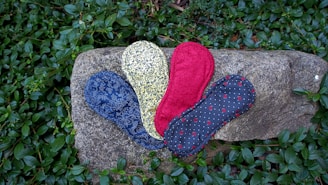 A collection of patterned reusable cloth pads are arranged on a large stone surrounded by green foliage. The pads feature different designs: one is blue with intricate floral patterns, another is yellow with black floral motifs, one is red with a solid color, and another is dark gray with small red details and white dots.