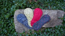 A collection of patterned reusable cloth pads are arranged on a large stone surrounded by green foliage. The pads feature different designs: one is blue with intricate floral patterns, another is yellow with black floral motifs, one is red with a solid color, and another is dark gray with small red details and white dots.