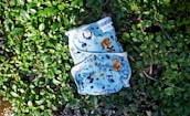 A cloth diaper with colorful animal and nature illustrations rests on a bed of lush green leaves. The illustrations include owls, raccoons, and various trees and plants, all set against a light blue background.