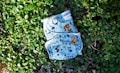 A cloth diaper with colorful animal and nature illustrations rests on a bed of lush green leaves. The illustrations include owls, raccoons, and various trees and plants, all set against a light blue background.