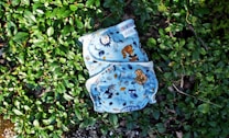 A cloth diaper with colorful animal and nature illustrations rests on a bed of lush green leaves. The illustrations include owls, raccoons, and various trees and plants, all set against a light blue background.