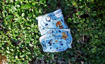 A cloth diaper with colorful animal and nature illustrations rests on a bed of lush green leaves. The illustrations include owls, raccoons, and various trees and plants, all set against a light blue background.