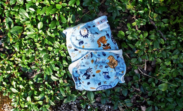 A cloth diaper with colorful animal and nature illustrations rests on a bed of lush green leaves. The illustrations include owls, raccoons, and various trees and plants, all set against a light blue background.