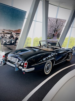 A classic black convertible car is parked inside a showroom with large windows. Large photographs hang in the background, depicting vintage racing cars and a scene with a woman near blossoming trees. The car has a stylish design with white wall tires and chrome accents.
