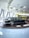 A classic black limousine is displayed on a round platform inside a modern showroom with large windows. The interior features a futuristic ceiling design with recessed circular lights. In the background, a bright yellow bus with visible signage is parked.