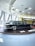 A classic black limousine is displayed on a round platform inside a modern showroom with large windows. The interior features a futuristic ceiling design with recessed circular lights. In the background, a bright yellow bus with visible signage is parked.