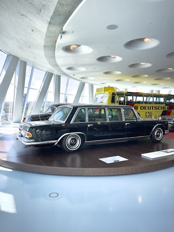 A classic black limousine is displayed on a round platform inside a modern showroom with large windows. The interior features a futuristic ceiling design with recessed circular lights. In the background, a bright yellow bus with visible signage is parked.