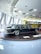 A classic black limousine is displayed on a round platform inside a modern showroom with large windows. The interior features a futuristic ceiling design with recessed circular lights. In the background, a bright yellow bus with visible signage is parked.