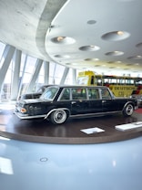 A classic black limousine is displayed on a round platform inside a modern showroom with large windows. The interior features a futuristic ceiling design with recessed circular lights. In the background, a bright yellow bus with visible signage is parked.