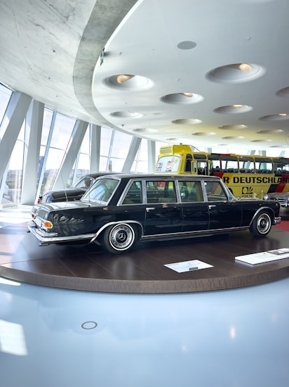 A classic black limousine is displayed on a round platform inside a modern showroom with large windows. The interior features a futuristic ceiling design with recessed circular lights. In the background, a bright yellow bus with visible signage is parked.