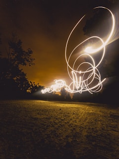 A long exposure photograph capturing swirling light trails against a dark night sky. The ground appears textured and earthy beneath the glowing patterns. Silhouetted trees frame the luminous display, adding depth and contrast.