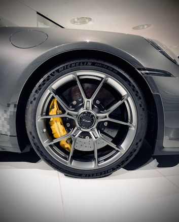 A close-up view of a sleek, modern car tire with a detailed alloy wheel design. The wheel features a vibrant yellow brake caliper visible through the spokes, while the tire brand and specifications are embossed along the sidewall.