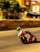 A motorcyclist in colorful racing gear leans into a sharp turn during a high-speed street race. The blurred background suggests motion, with hay bales serving as a barrier on the side of the road. Buildings and trees line the street, adding an urban feel to the race setting.