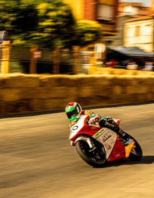 A motorcyclist in colorful racing gear leans into a sharp turn during a high-speed street race. The blurred background suggests motion, with hay bales serving as a barrier on the side of the road. Buildings and trees line the street, adding an urban feel to the race setting.