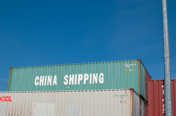 Three stacked shipping containers are visible, with the top one labeled 'China Shipping' in bold white letters. A clear blue sky serves as the background, and there is a vertical metal pole on the right side of the image.