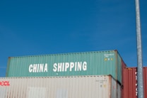 Three stacked shipping containers are visible, with the top one labeled 'China Shipping' in bold white letters. A clear blue sky serves as the background, and there is a vertical metal pole on the right side of the image.