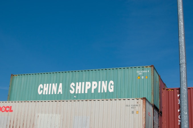Three stacked shipping containers are visible, with the top one labeled 'China Shipping' in bold white letters. A clear blue sky serves as the background, and there is a vertical metal pole on the right side of the image.