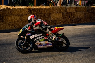 A motorcyclist in full racing gear is leaning into a turn on a track lined with hay bales. The background features a barrier and some foliage. The motorcycle is covered with various sponsor decals and features a vibrant design.