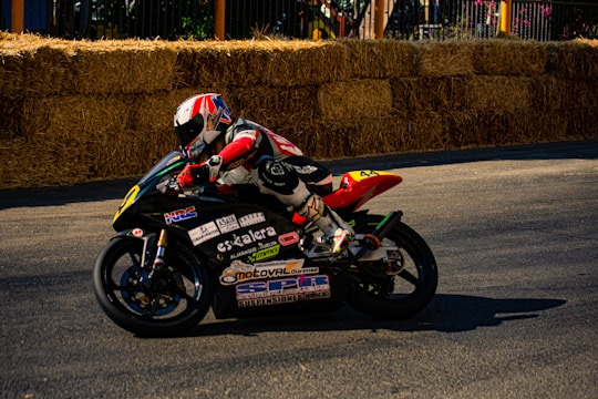 A motorcyclist in full racing gear is leaning into a turn on a track lined with hay bales. The background features a barrier and some foliage. The motorcycle is covered with various sponsor decals and features a vibrant design.