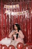 A young woman wearing a sash and a tiara is posing in front of a red tinsel backdrop. Above her, a banner spells out 'Happy Birthday' in golden letters. She is making a heart shape with her hands, and there are several red balloons in the foreground along with a fluffy white cushion that has a small birthday cake on it.