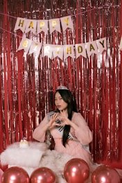 A young woman wearing a sash and a tiara is posing in front of a red tinsel backdrop. Above her, a banner spells out 'Happy Birthday' in golden letters. She is making a heart shape with her hands, and there are several red balloons in the foreground along with a fluffy white cushion that has a small birthday cake on it.