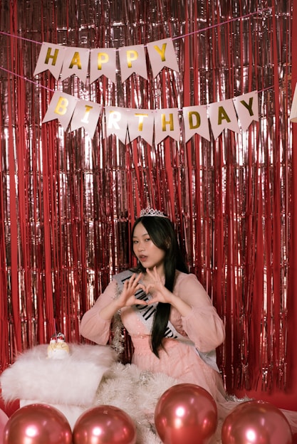 A young woman wearing a sash and a tiara is posing in front of a red tinsel backdrop. Above her, a banner spells out 'Happy Birthday' in golden letters. She is making a heart shape with her hands, and there are several red balloons in the foreground along with a fluffy white cushion that has a small birthday cake on it.