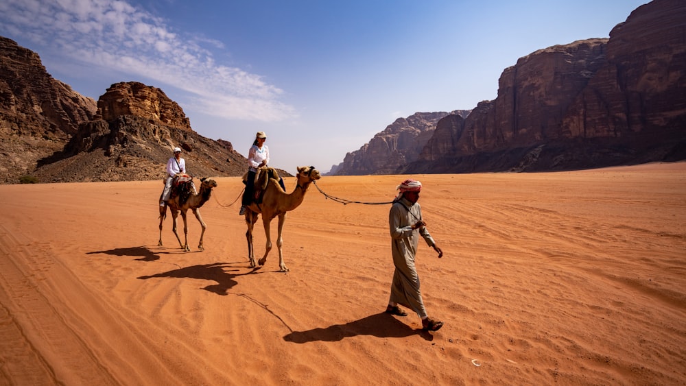 a person walking with a group of camels on a sandy hill with Wadi Rum in the background