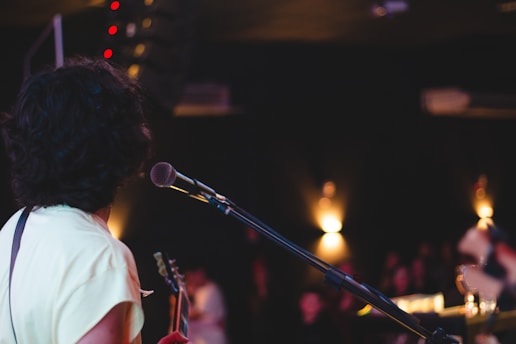 A musician with curly hair stands on stage, holding a guitar and facing away from the camera. A microphone is positioned in front of them. The background is dimly lit, with blurred lights and silhouettes of audience members.