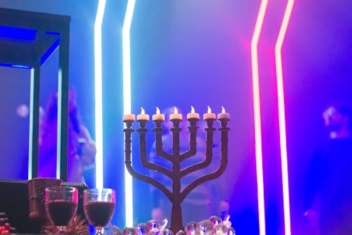 A menorah with lit candles stands prominently against a backdrop of vibrant LED lights in blue and pink. In the foreground, a couple of wine glasses are partially visible, adding a festive touch. The setting appears to be a dimly lit indoor space, possibly for a celebration or event.