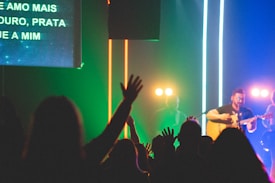 A lively music concert scene with a singer playing a guitar on stage. The audience is engaged, with many people raising their hands in the foreground. Bright stage lights and colorful neon stripes add a vibrant atmosphere. Partial view of lyrics is visible on a screen at the left.