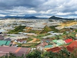 A vast landscape features numerous greenhouses spread across a hilly area. The scene includes small buildings with colorful roofs, surrounded by green vegetation and manicured agricultural plots. In the background, mountains are visible under a sky filled with clouds.