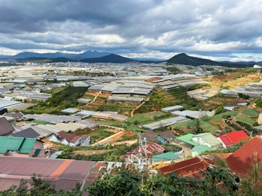 A vast landscape features numerous greenhouses spread across a hilly area. The scene includes small buildings with colorful roofs, surrounded by green vegetation and manicured agricultural plots. In the background, mountains are visible under a sky filled with clouds.