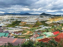 A vast landscape features numerous greenhouses spread across a hilly area. The scene includes small buildings with colorful roofs, surrounded by green vegetation and manicured agricultural plots. In the background, mountains are visible under a sky filled with clouds.