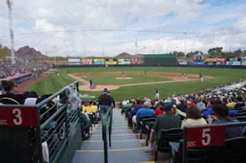 A baseball game is taking place in a stadium with a sizeable audience in the stands. The field is well-maintained, with players positioned strategically for the game. The audience appears engaged, some wearing team colors. In the background, there are billboard advertisements and a scenic view of distant mountains under a partly cloudy sky.