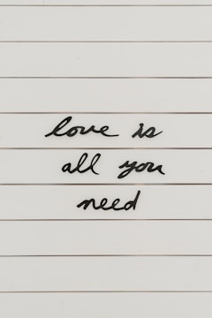 Black cursive letters spell out the phrase 'love is all you need' on a white, lined background.