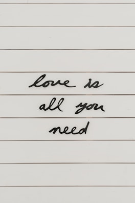 Black cursive letters spell out the phrase 'love is all you need' on a white, lined background.