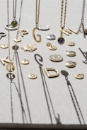 A variety of jewelry pieces, including necklaces with letter pendants and round charms, are displayed on a textured surface. The letters are of varying styles and sizes, suspended among other small, intricate decorative items. Shadows cast by the jewelry create an artistic pattern on the surface.