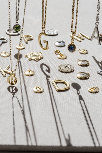 A variety of jewelry pieces, including necklaces with letter pendants and round charms, are displayed on a textured surface. The letters are of varying styles and sizes, suspended among other small, intricate decorative items. Shadows cast by the jewelry create an artistic pattern on the surface.