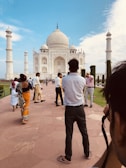 A group of people walking on a pathway toward a large white marble mausoleum with a central dome and four minarets. The sky above is clear with some clouds, and there are trees and greenery visible in the vicinity. The scene is lively with visitors of different ages and styles of dress.