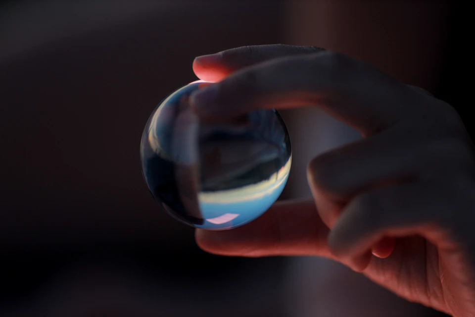 The image shows a hand holding a small, round glass ball with a blue and white patterned surface against a dark background. The hand is positioned on the left side of the frame, while the glass ball is located on the right side.