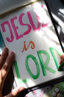 A hand-drawn illustration features the words 'JESUS is LORD' written in large, colorful letters on a sheet of paper. The word 'JESUS' is in pink, 'is' is in orange, and 'LORD' is in green. The paper is resting on a surface that appears to be covered in artistic materials, hinting at an artistic setting. A hand is visible, possibly holding a green marker.