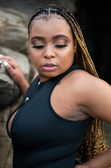 A woman with braided hair wearing a black dress poses with her eyes gently closed. Her expression is serene, and she stands against a rock backdrop.