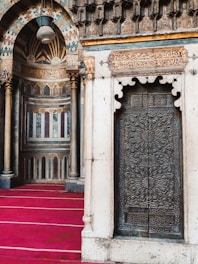 A richly ornamented interior features intricate geometric and floral patterns on a metal door, framed by stone architecture. The wall displays elaborate Arabic calligraphy in golden hues, accentuated by cylindrical columns. The flooring is covered with a deep red carpet, adding warmth and depth to the space.