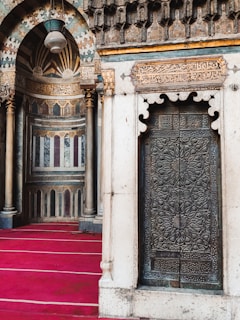 A richly ornamented interior features intricate geometric and floral patterns on a metal door, framed by stone architecture. The wall displays elaborate Arabic calligraphy in golden hues, accentuated by cylindrical columns. The flooring is covered with a deep red carpet, adding warmth and depth to the space.