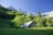 A rustic wooden cabin with a tin roof is nestled in a lush, green meadow surrounded by dense forest. In the background, towering snow-capped mountains rise majestically against a clear blue sky.
