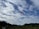 A single paraglider with an orange parachute is soaring in the sky above a scenic landscape. The sky is filled with a mix of fluffy white clouds against a deep blue background. Below, lush green hills and fields stretch out, adding a sense of tranquility and openness to the scene.