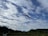 A single paraglider with an orange parachute is soaring in the sky above a scenic landscape. The sky is filled with a mix of fluffy white clouds against a deep blue background. Below, lush green hills and fields stretch out, adding a sense of tranquility and openness to the scene.